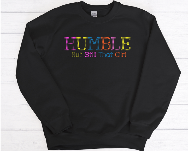 Black sweatshirt with 'HUMBLE But Still That Girl' text on a white wooden background